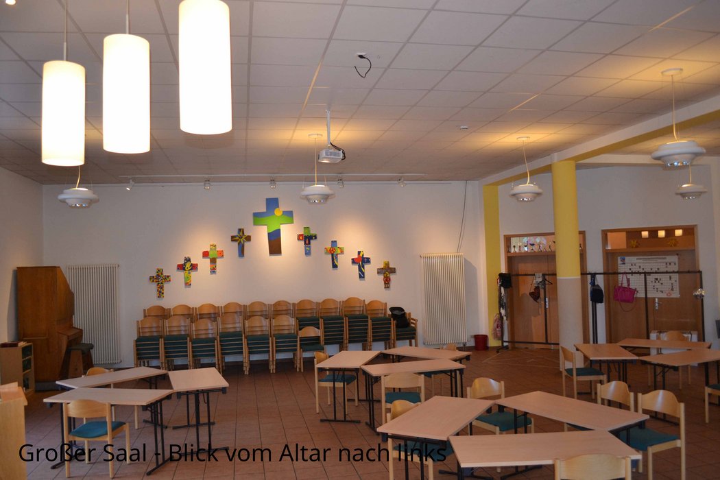 Das Foto zeigt einen Blick über Tische und Stühle hinweg in den Raum hinein. An der gegenüberliegenden Seite befinden sich ein großes und acht kleinere Kreuze an der Wand, die in einer Aktion mit Farben gestaltet wurden