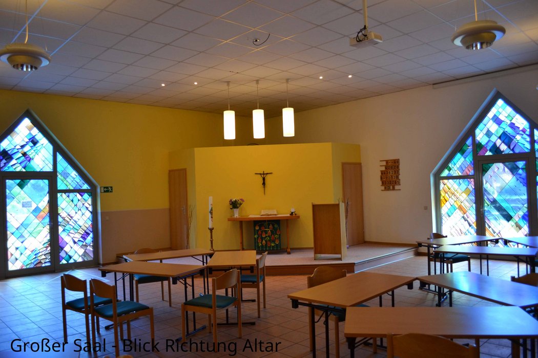Blick auf den Altar. Rechts und links wird der Altar von bunten Fenstern eingerahmt. An der Wand hängt ein Kreuz, über dem Alter leuchten drei Hängelampen.