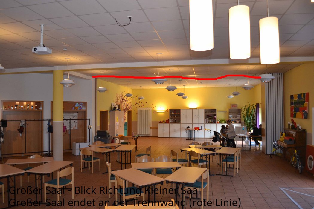 Das Foto zeigt einen Blick über Tische und Stühle hinweg in den Raum hinein. Durch eine rote Linie soll gezeigt werden, wo eine aus mehreren Teilen bestehende Zwischenwand eingezogen werden kann. An der gegenüberliegenden Seite befinden sich mehrere Materialschränke.